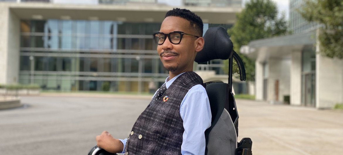 A South African man in a wheelchair and glasses, wearing a vest, smiles in front of a modern building.