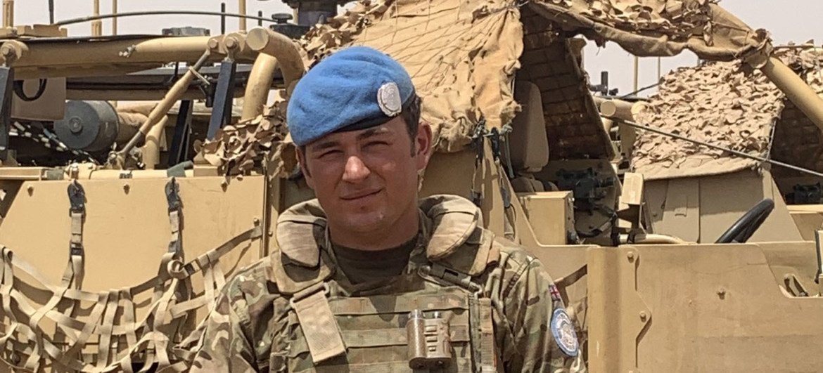 British soldier Jack Drake in uniform and blue beret, standing in front of an armored vehicle during a UN peacekeeping mission in Mali.